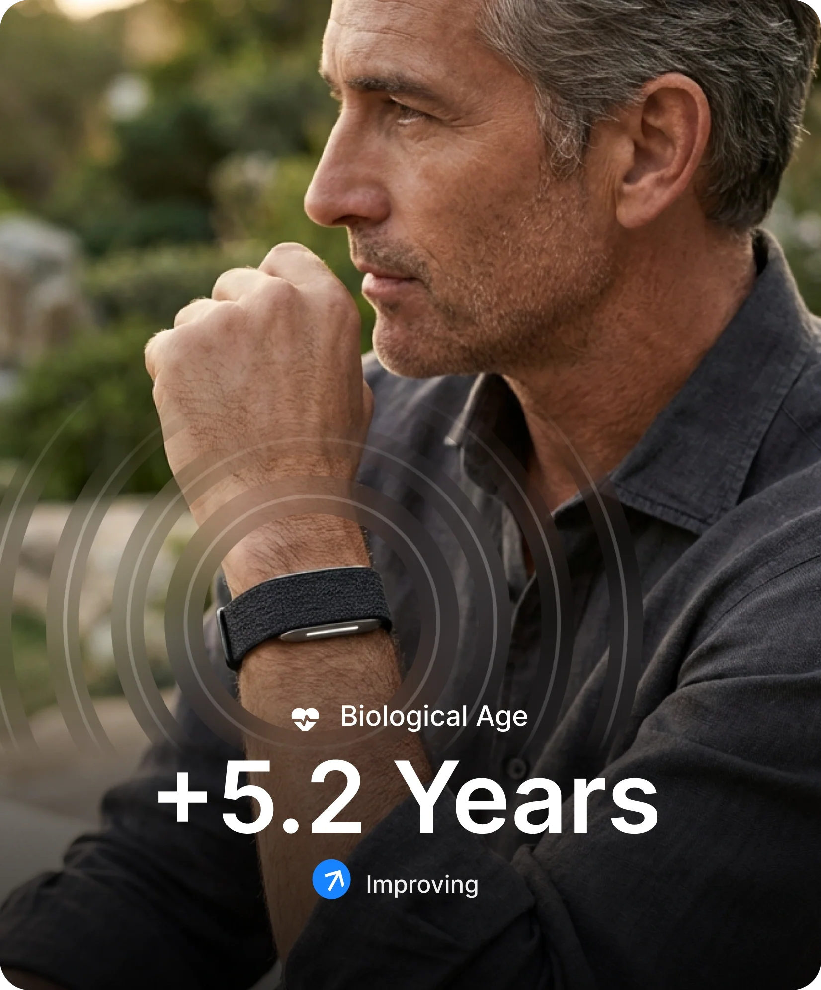 Middle-aged person wearing a hume band 2.0 while seated outdoors. A digital overlay displays a Biological Age score of +5.2 years with an improving trend indicator.